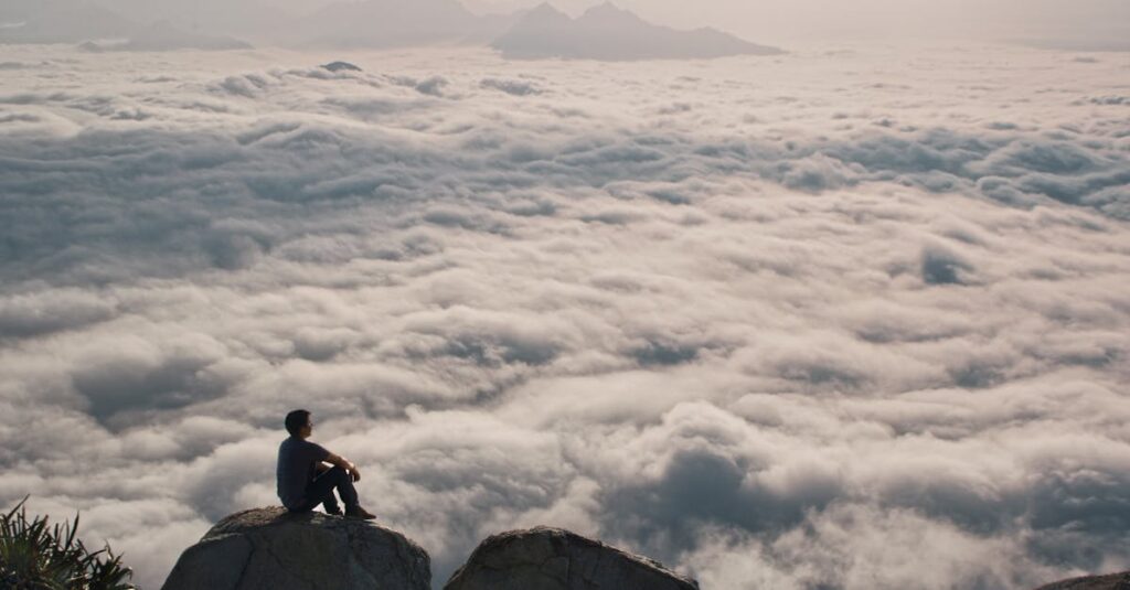 Person sitting on a mountain peak above clouds, capturing serenity and adventure.